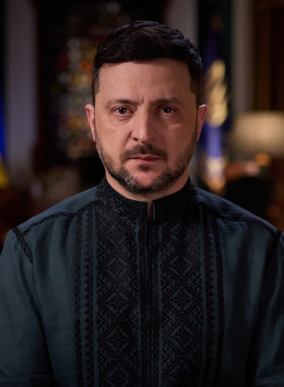 Volodymyr Zelenskyj i mörk broderad skjorta blickar allvarligt in i kameran, med ukrainska flaggor suddiga i bakgrunden.