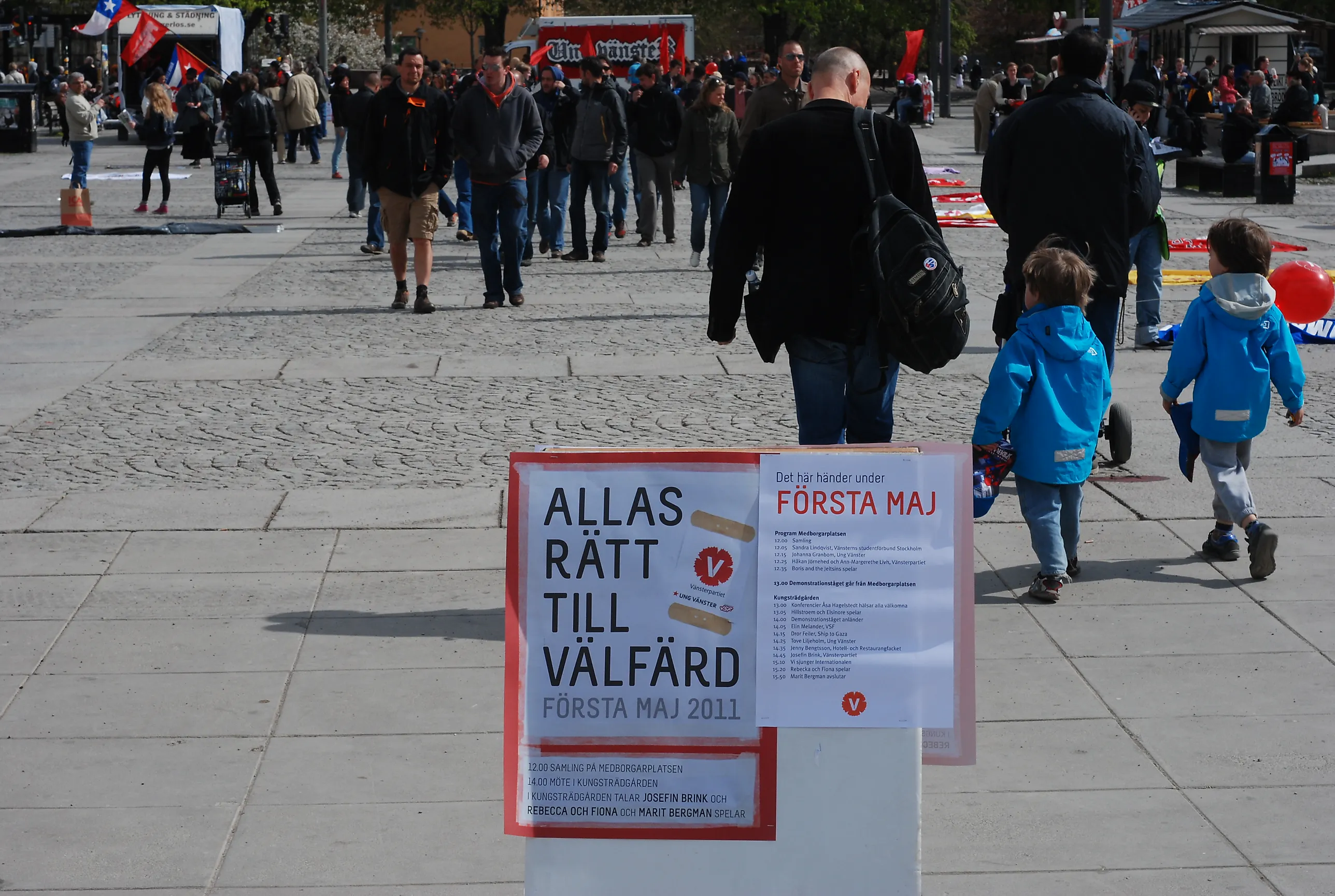 En affisch med texten "Allas rätt till välfärd" och Vänsterpartiets logotyp står på ett torg där människor rör sig i bakgrunden.