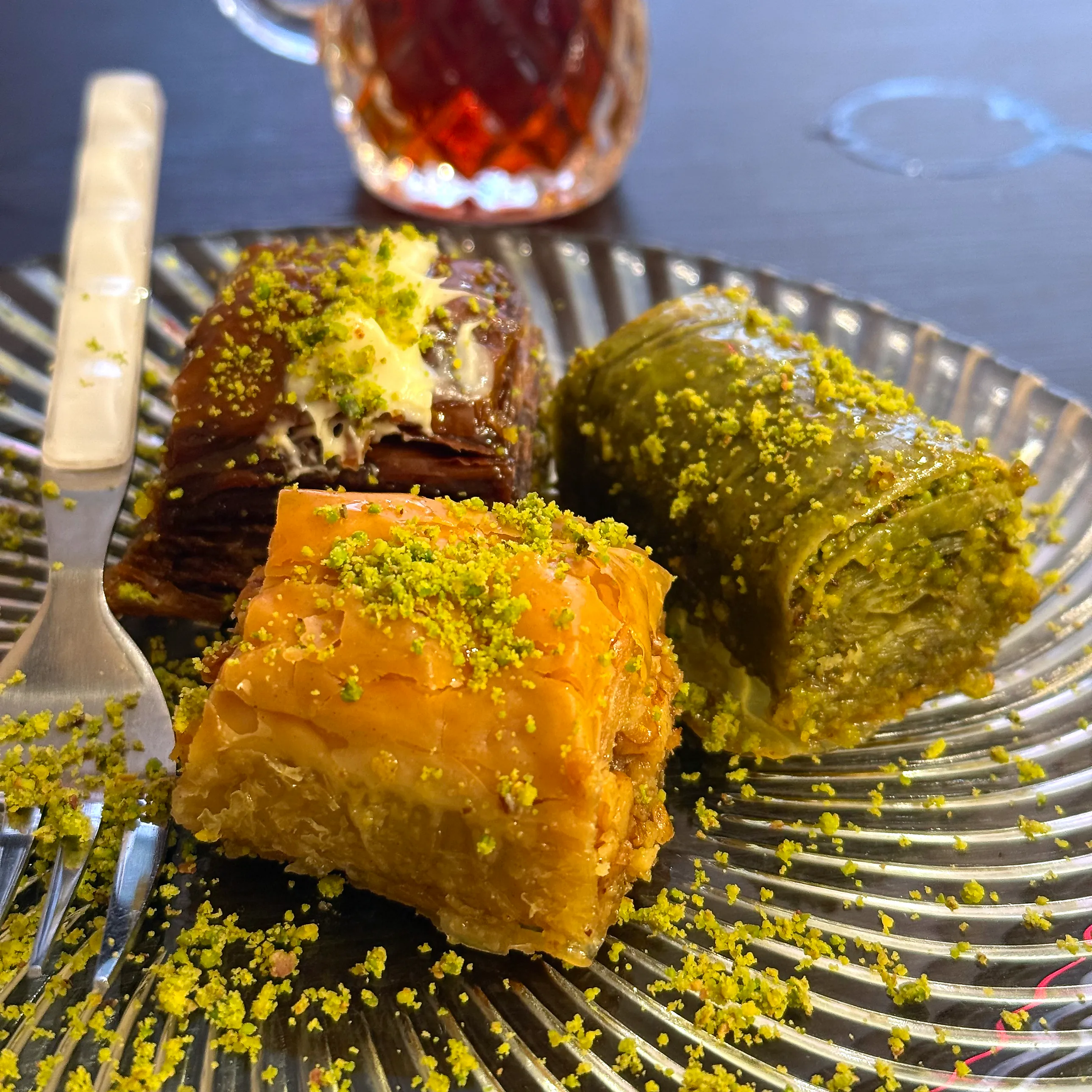 Tre bitar baklava på ett räfflat glasfat – en gyllene fyrkant, en mörk chokladbit med krämig fyllning och en rulle med grön pistagefyllning – alla rikligt strödda med malet pistage, med gaffel och ett glas svart te intill.