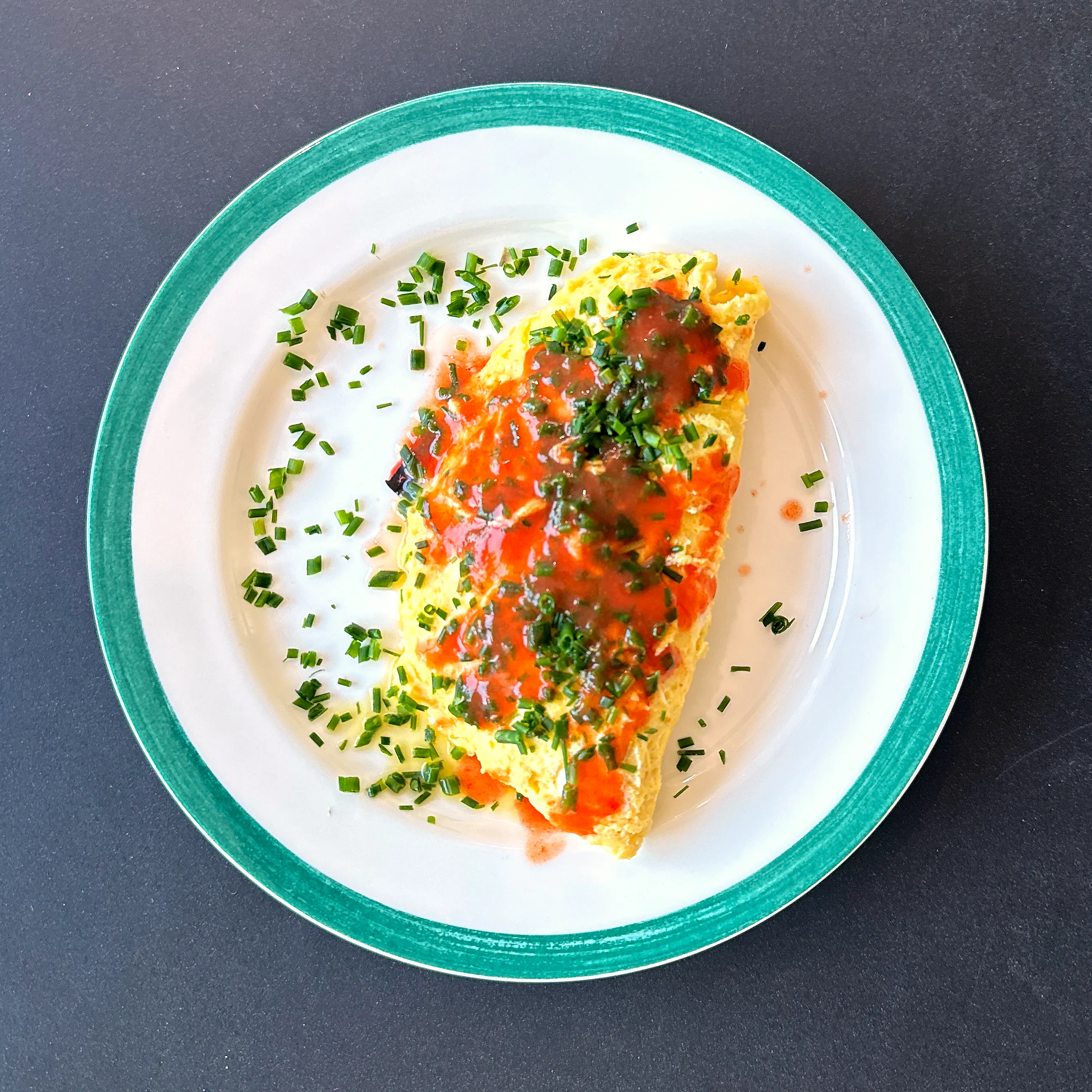 En hopvikt gul omelett på en vit tallrik med grön kant, ringlad med röd chilisås och strödd med finklippt gräslök som även ligger på tallrikskanten.