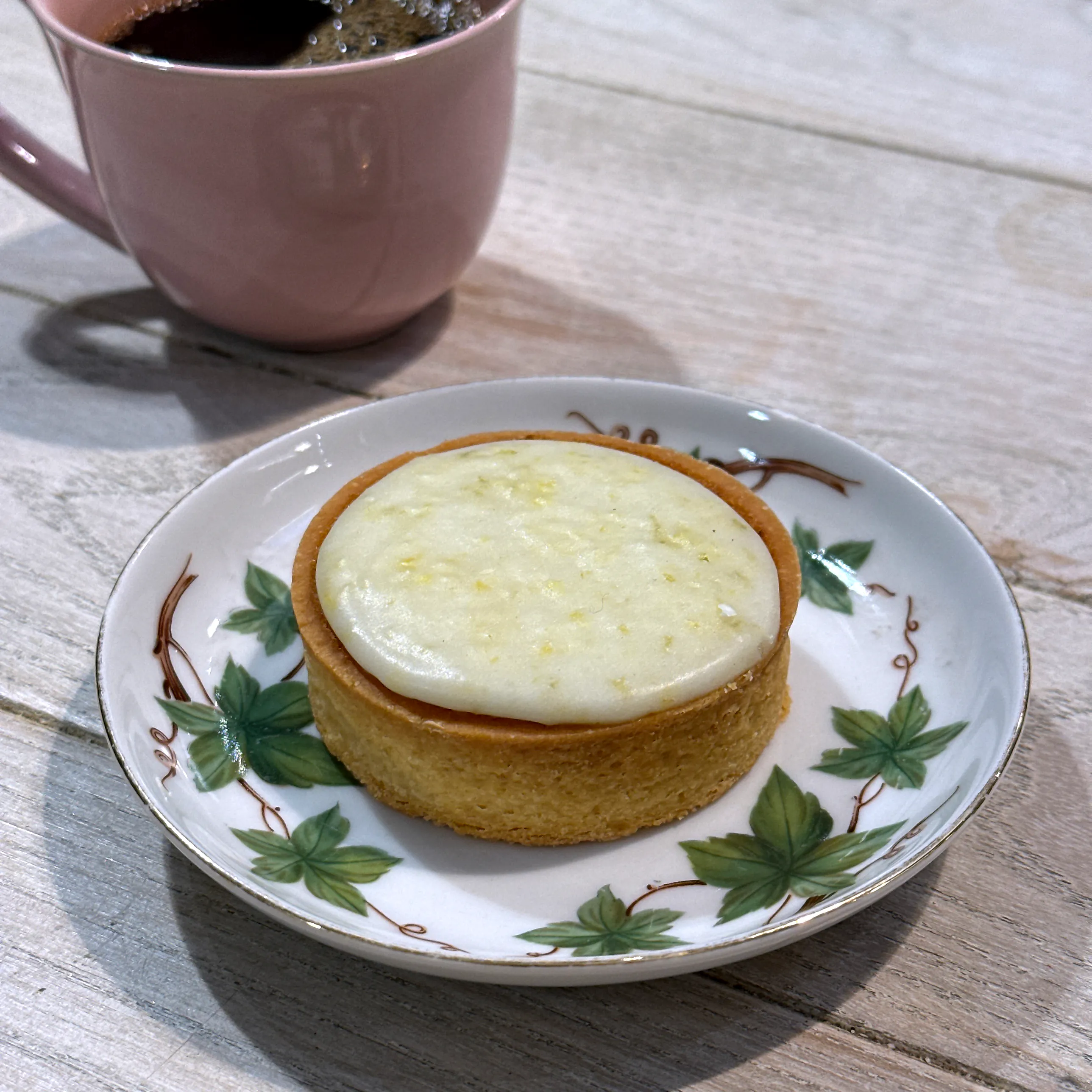 En rund mazarin med gyllene mördegskant och blank, ljus citronglasyr prickad av rivet citronskal, på ett vitt fat med murgrönsmönster intill en rosa kaffekopp.