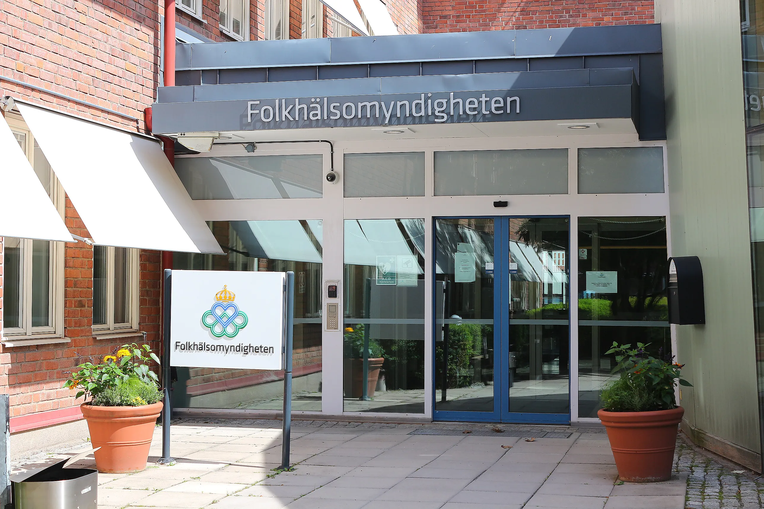 Entréparti med skjutdörrar i glas och blå ram, skylt med Folkhälsomyndighetens namn och logotyp framför en tegelbyggnad.
