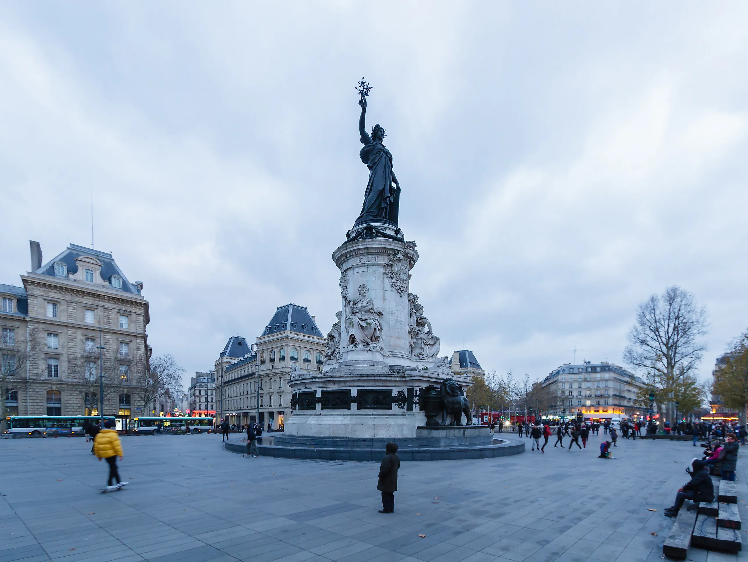 Ett stort öppet torg med gatsten och enstaka förbipasserande, framför ett högt stenmonument krönt av en bronsstatyk av Marianne, omgiven av parisiska stenhus en grå vinterdag.