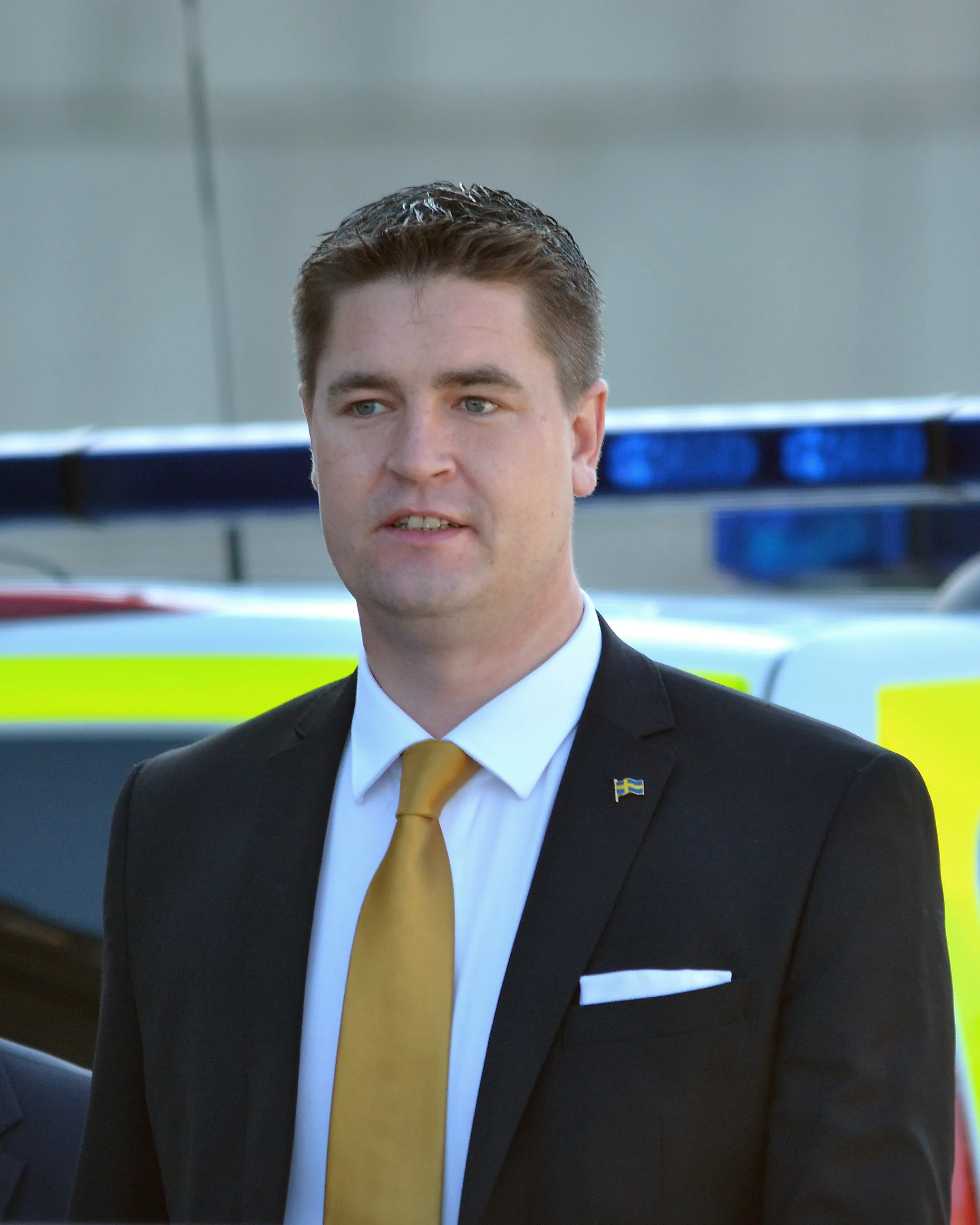 Oscar Sjöstedt i mörk kostym och gul slips med en svensk flagga i kavajslaget, framför polisbilar.